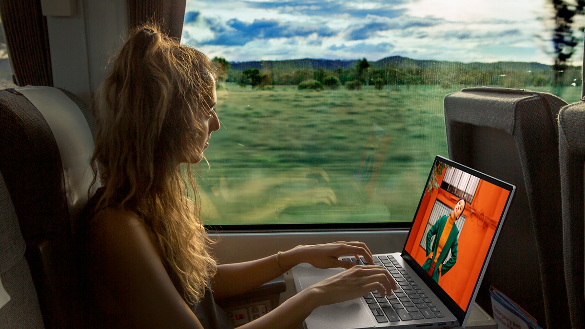 Image d’une femme dans un train, utilisant un ordinateur portable Dell sur la table basse face à elle. Une grande fenêtre dévoile le paysage.