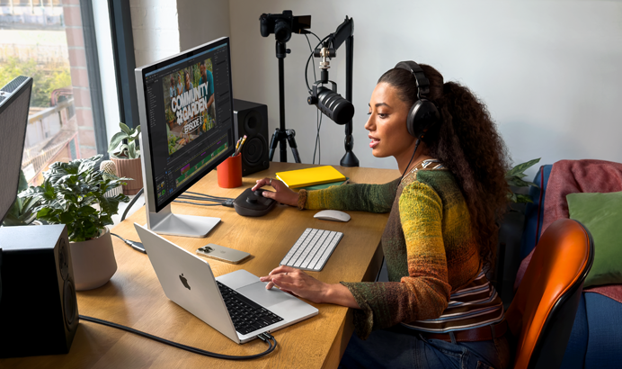 Une femme chez elle, travaillant à son bureau sur un MacBook Pro connecté à deux écrans externes, portant un casque et parlant dans un micro