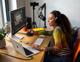 Une femme chez elle, travaillant à son bureau sur un MacBook Pro connecté à deux écrans externes, portant un casque et parlant dans un micro